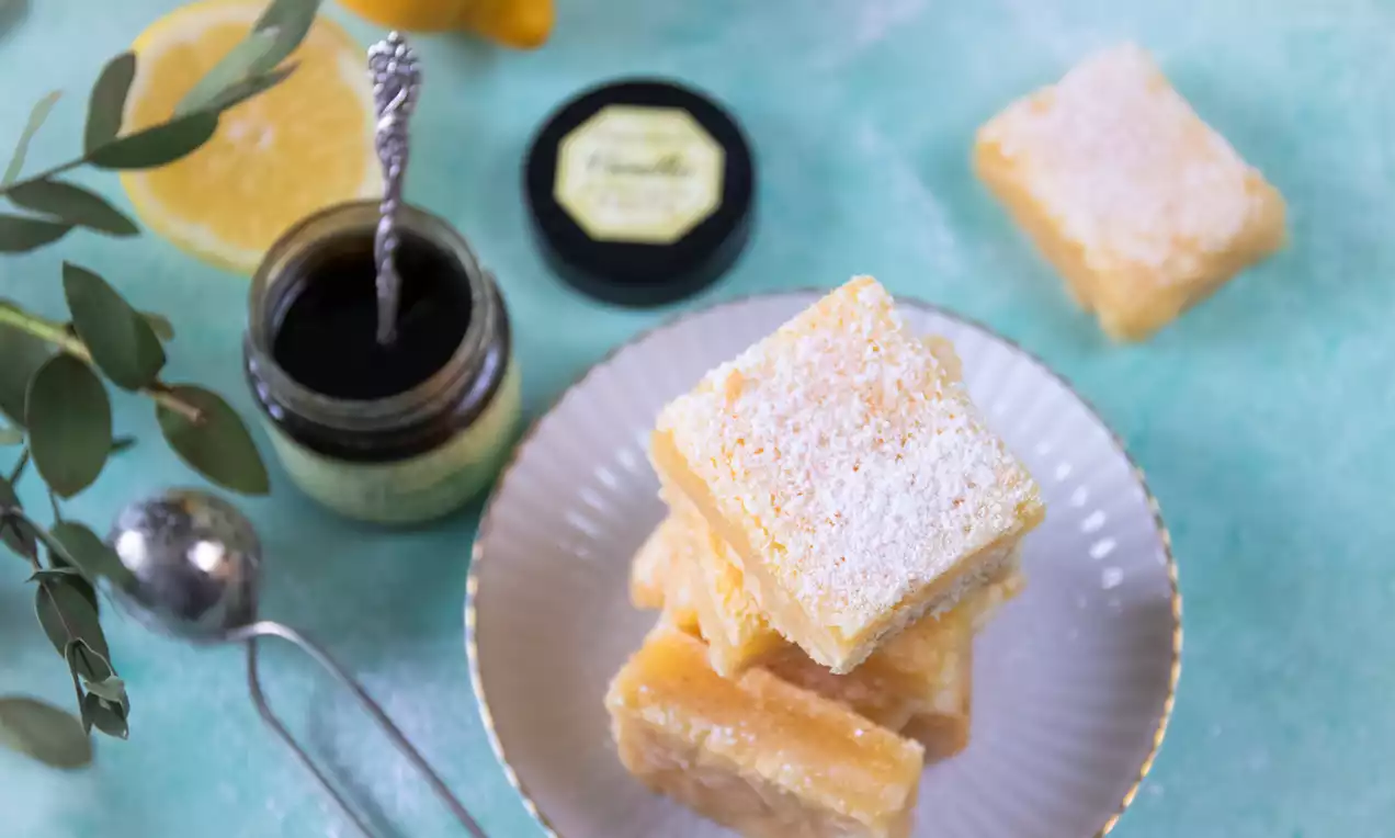 Citronrutor med gyllengul fyllning och pudrat florsocker staplade på vit tallrik, omgivna av citronskiva, grön kvist och burk på turkos yta